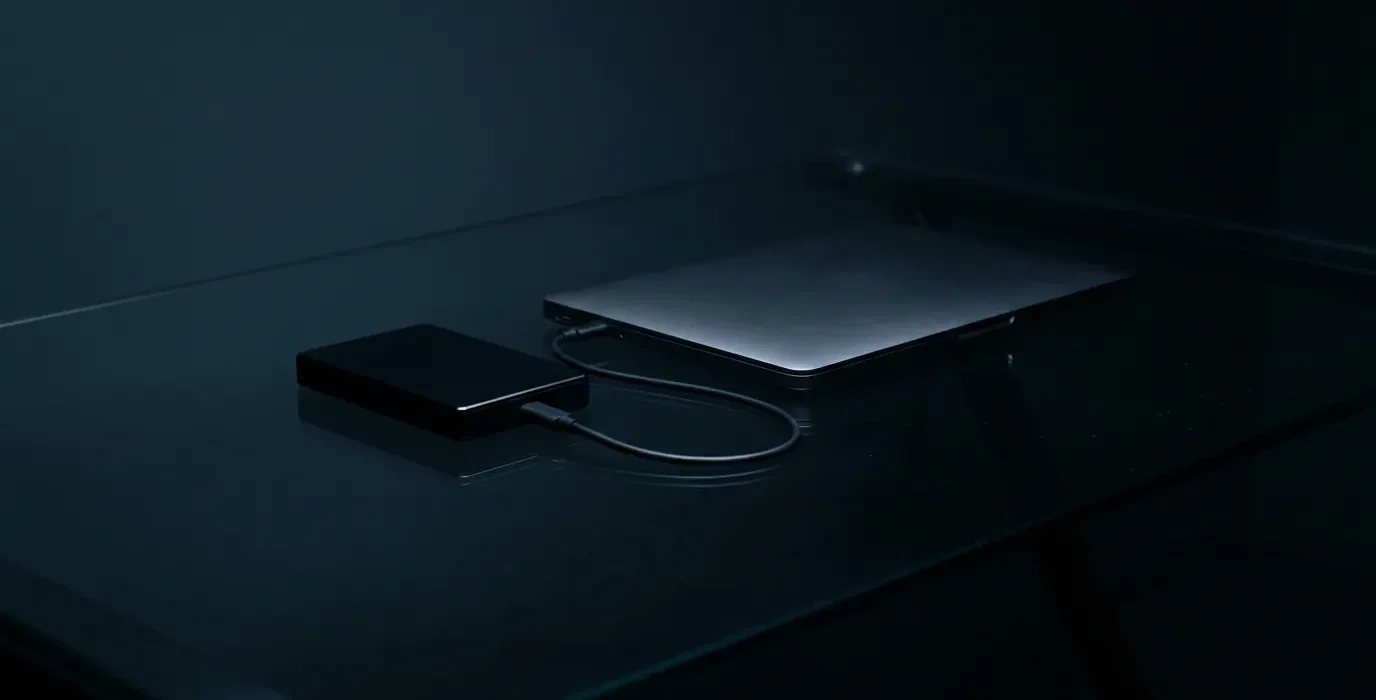 Un disque dur externe relié à un ordinateur portable fermé, sur un bureau en verre sombre