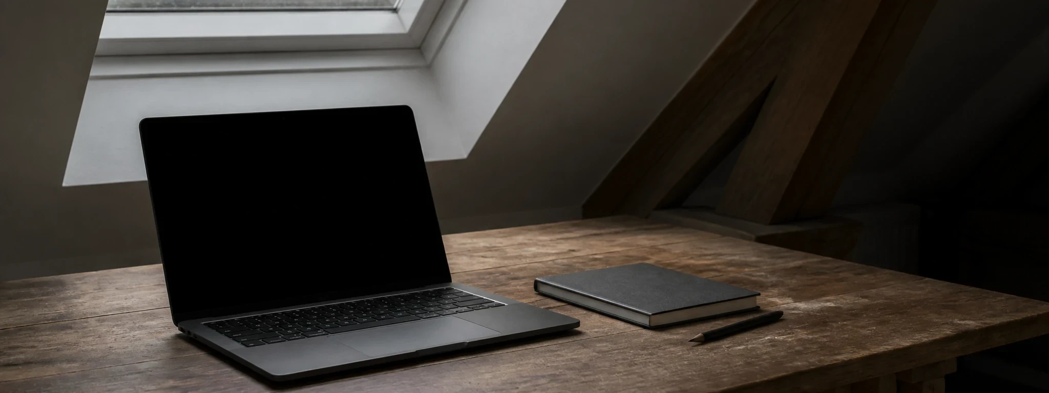 Un ordinateur portable ouvert sur un bureau en bois brut, sous la lumière grise d'une fenêtre de toit